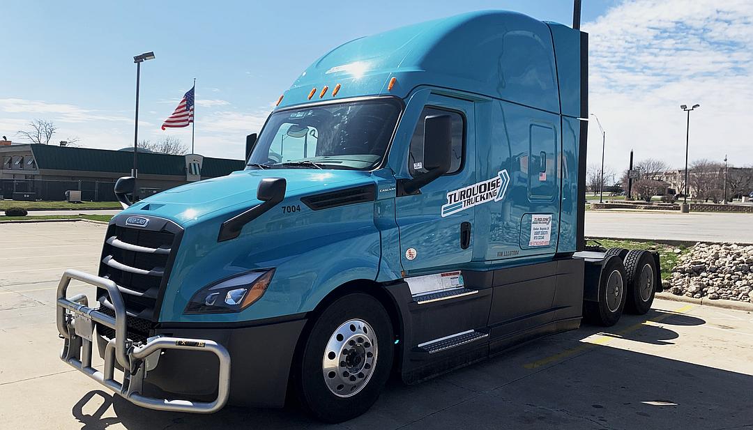 Blue Cascadia truck with Turquoise Trucking decals parked with an American flag behind it.